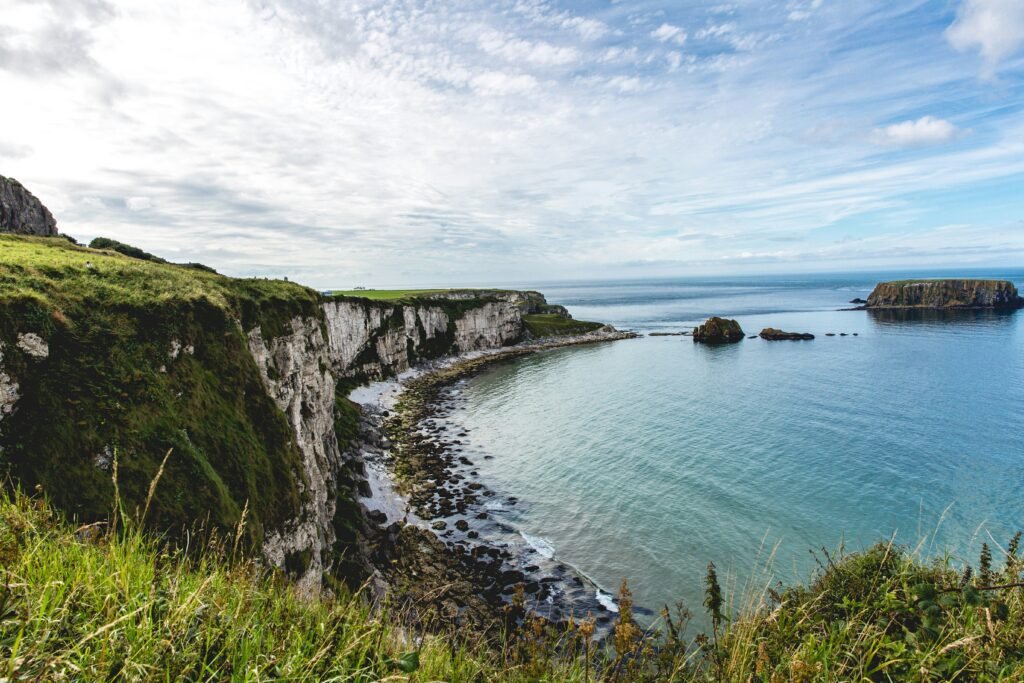 A scenic view of rugged cliffs and a calm bay along the Causeway Coastal Route in Ballintoy, Northern Ireland, with rocky islets in the distance.