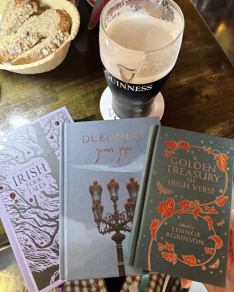 A table with three books: Dubliners by James Joyce, The Anthology of Irish Folk Tales, and A Golden Treasury of Irish Verse, alongside a pint of Guinness and a basket of Irish brown bread. This image showcases key Irish literature and culture, perfect for exploring what to read, watch, and listen to before visiting Ireland.