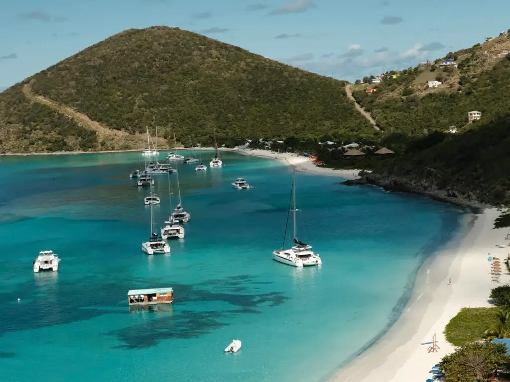 A stunning view of yachts anchored in clear blue waters along the white sandy beach of Jost Van Dyke in the British Virgin Islands, featuring lush green hills and a peaceful tropical atmosphere. A popular spot for visitors to the Soggy Dollar Bar.