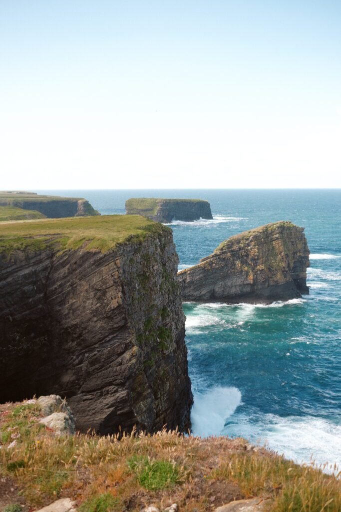 A breathtaking view of the Kilkee Cliffs in County Clare, Ireland, with dramatic, layered rock formations jutting out into the Atlantic Ocean. The turquoise waters crash against the cliff faces, and the grassy cliff tops add a vibrant contrast against the deep blue horizon. This hidden gem along Ireland’s Wild Atlantic Way offers stunning coastal scenery and is one of the best cliffs in Ireland to visit.