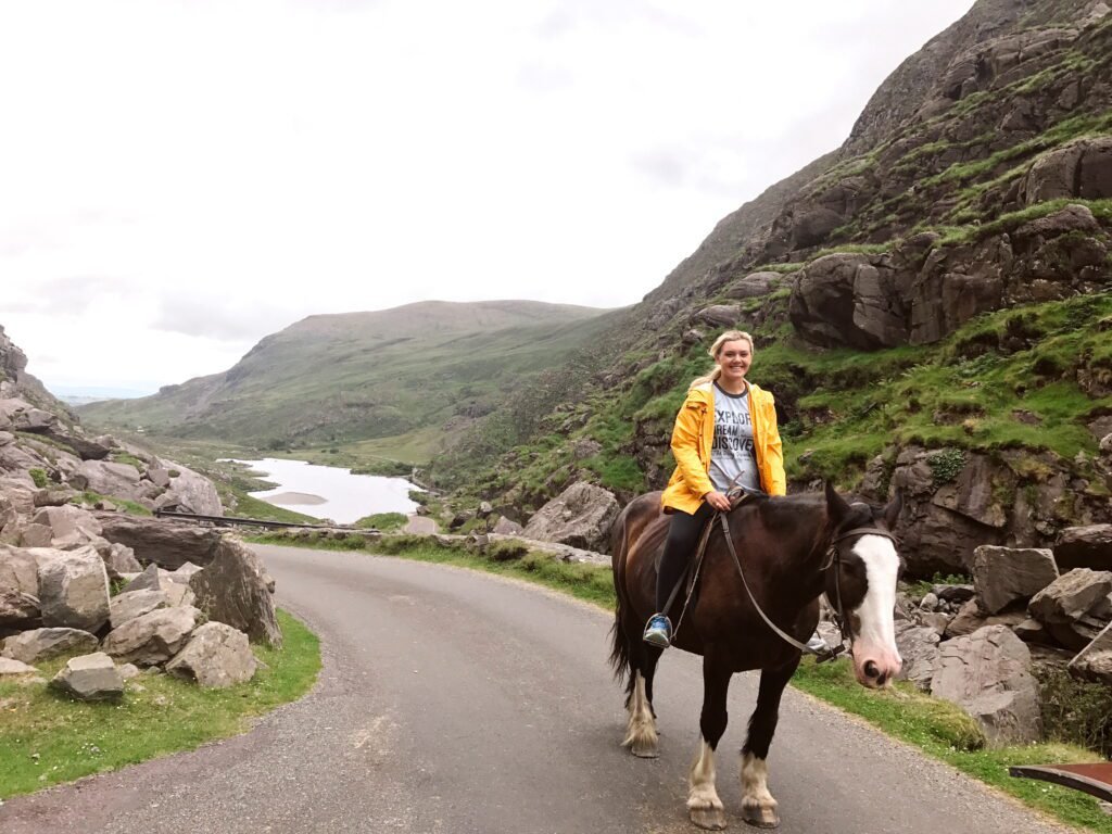 A traveler riding an Irish cob horse along a winding road in the Gap of Dunloe, surrounded by lush green mountains and overlooking a peaceful lake in the distance.