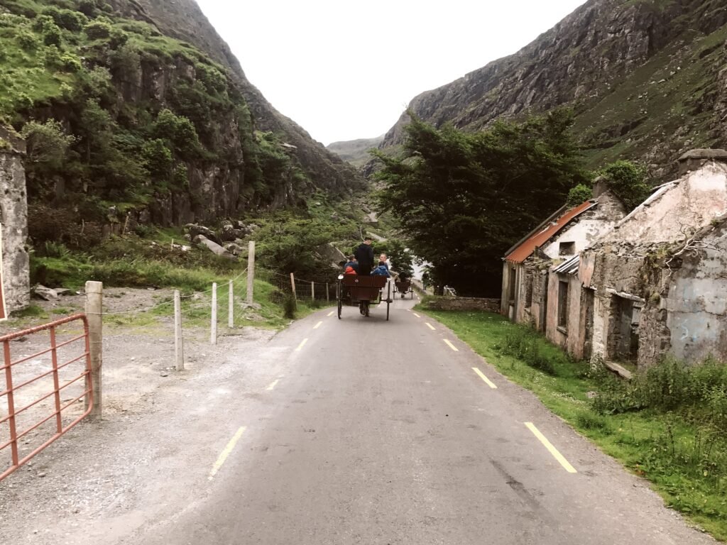 A jaunting car (horse-drawn carriage) travels along a narrow road through the Gap of Dunloe, surrounded by rugged mountains and old stone buildings.