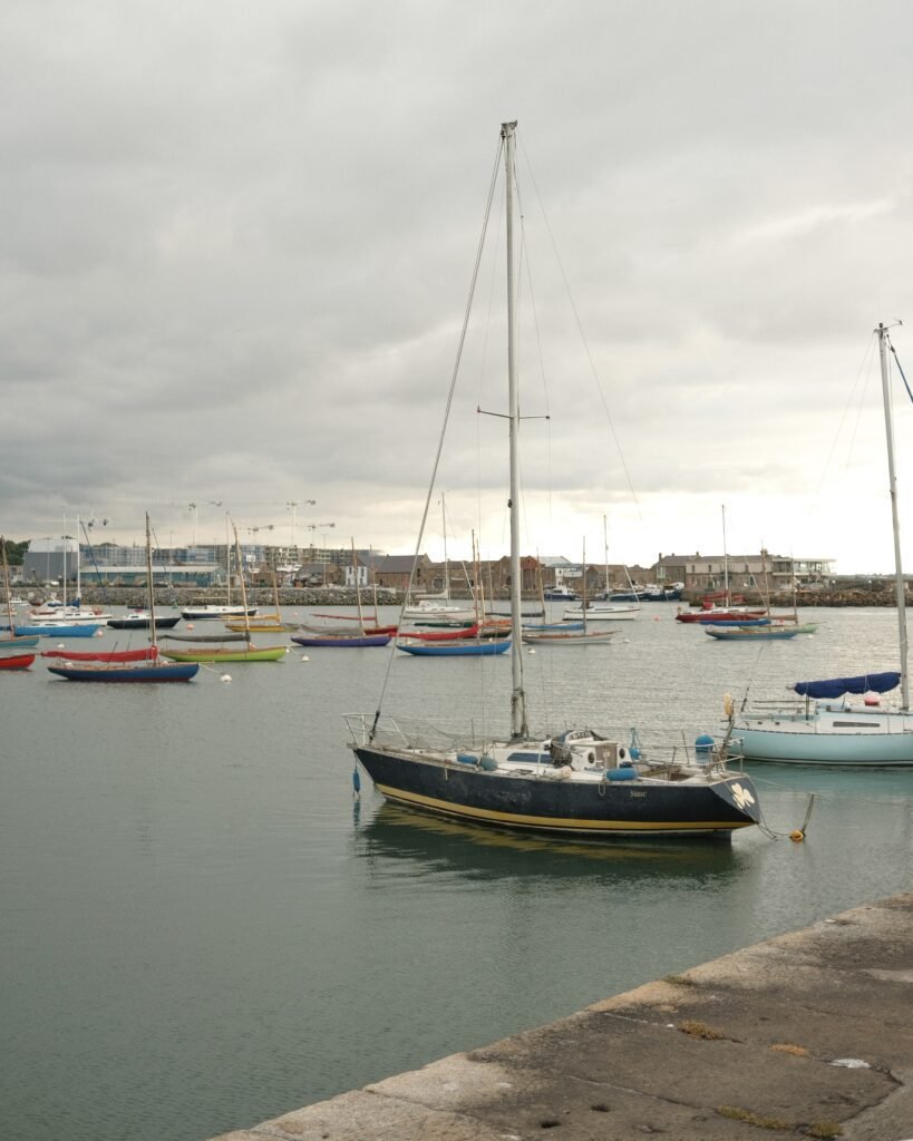 Sailboats anchored in the peaceful waters of Howth Harbor, Ireland, with cloudy skies and a seaside village in the background.