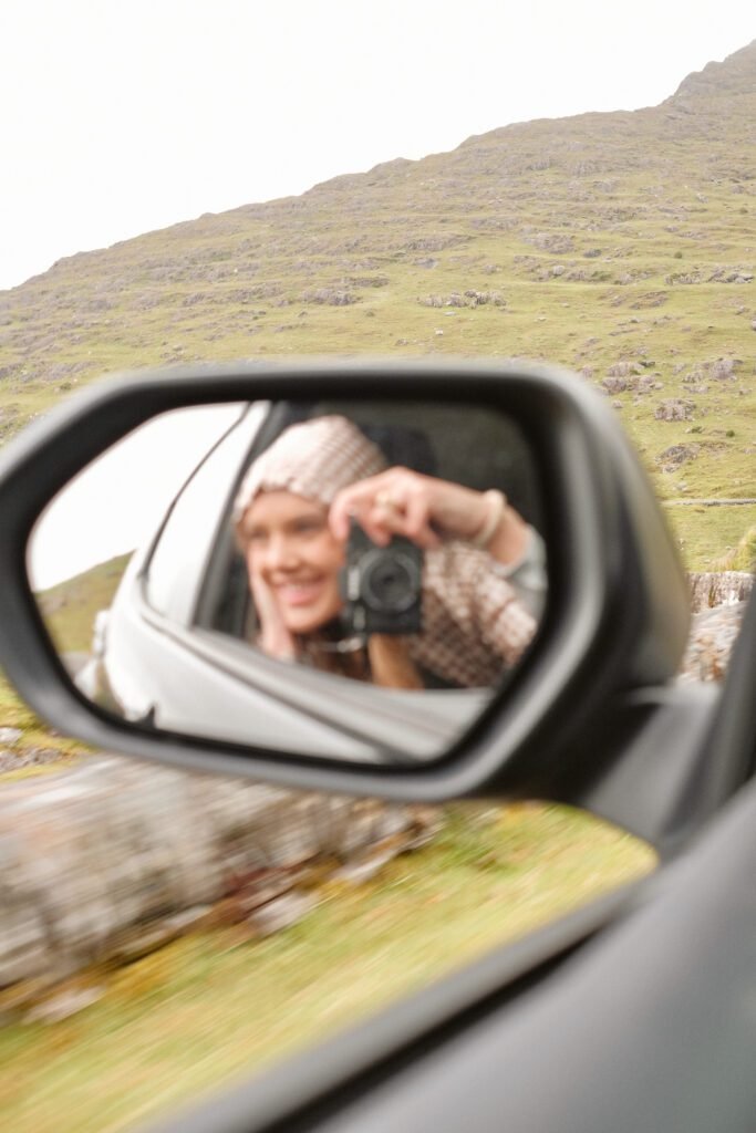 Kaley, travel blogger from Kindred Compass, captured in a car mirror taking photos during a road trip through Ireland.