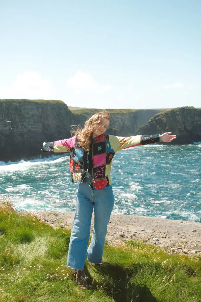 Kaley, travel blogger from Kindred Compass, standing at the Kilkee Cliffs in Ireland, arms outstretched with the ocean in the background.