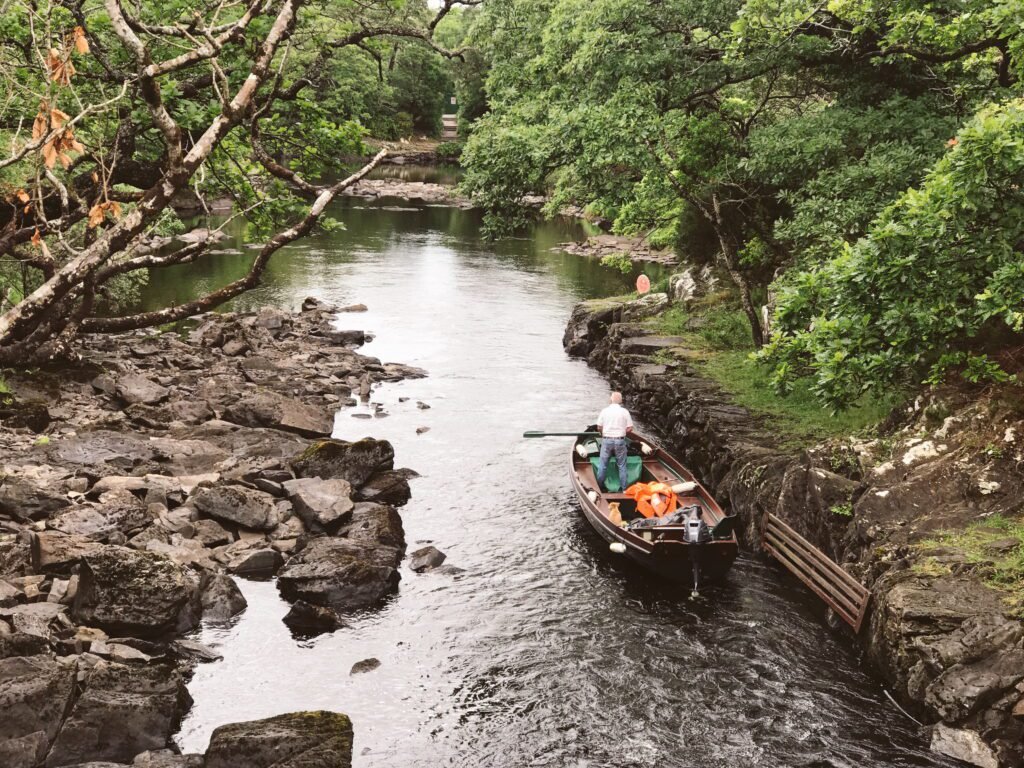 A boatman prepares a small boat for a ride through a narrow, rocky waterway surrounded by lush greenery in Killarney National Park.