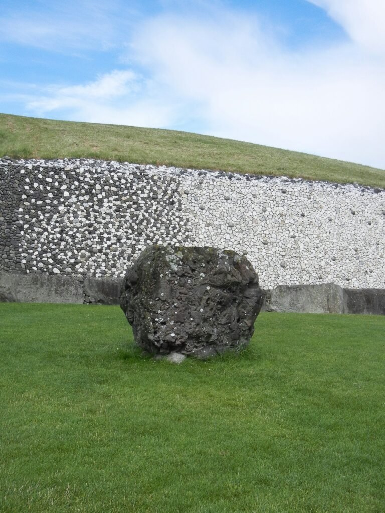 A large stone standing in front of the white stone wall of Newgrange, an ancient passage tomb in Ireland, with a green grass field and a blue sky.