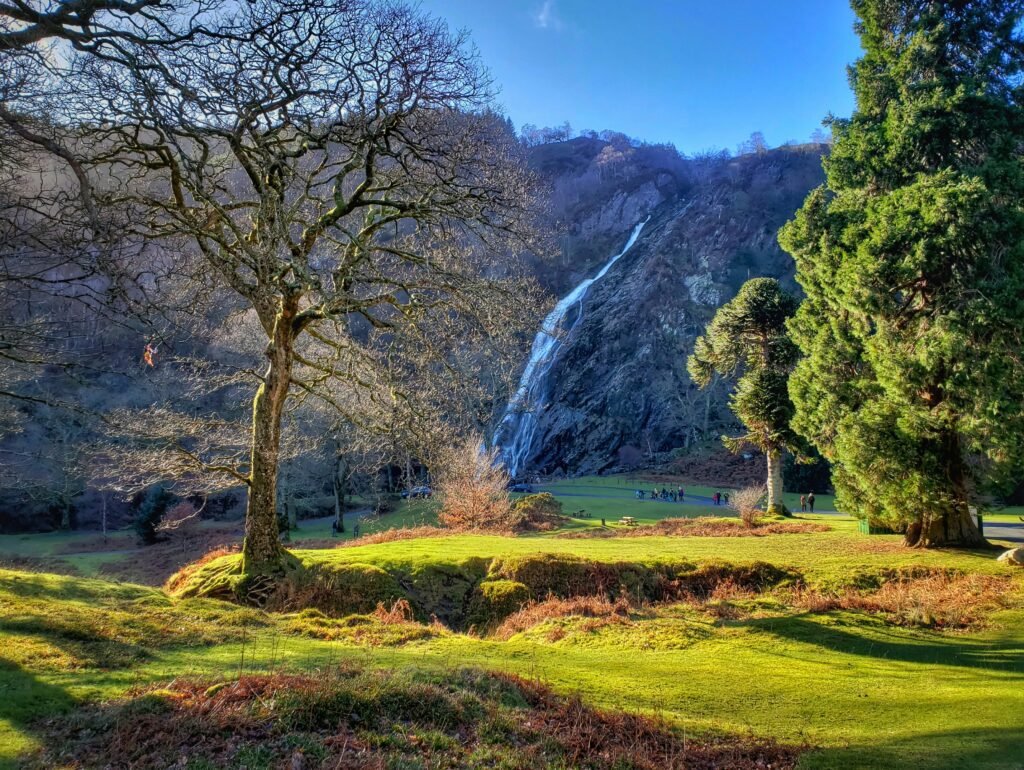 A majestic waterfall cascading down a rocky cliff, surrounded by lush trees and a grassy landscape at Powerscourt Waterfall in County Wicklow, Ireland.