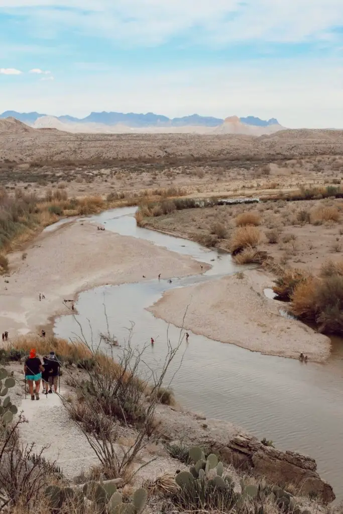 Hikers walking along the Rio Grande River near Big Bend National Park, with mountains and desert landscape in the background.