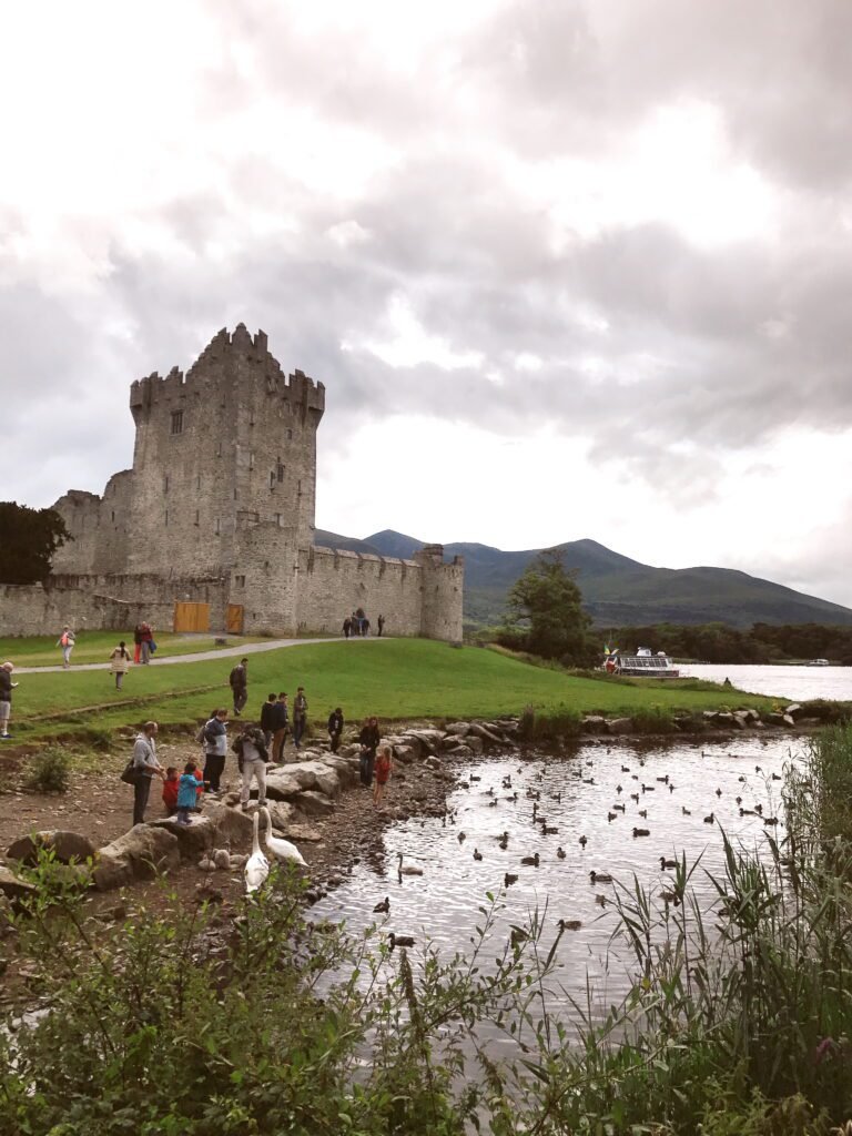 Visitors gather by the lakeshore near Ross Castle in Killarney National Park, with ducks swimming in the water and the castle’s stone tower overlooking the scene.