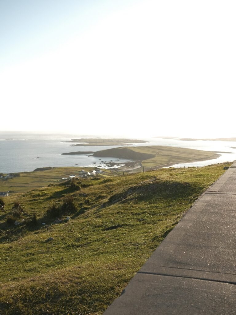 This image captures the breathtaking coastline view from The Sky Road in Clifden, part of the Wild Atlantic Way in Connemara, County Galway. The sunlit hills gently slope down towards the ocean, with scattered farmlands and cottages dotting the landscape. The road runs along the edge, offering stunning panoramic views of the Atlantic and the surrounding coastline, making it a must-visit for anyone looking to explore one of Ireland's most scenic drives. This peaceful and dramatic setting perfectly encapsulates the beauty of Connemara.