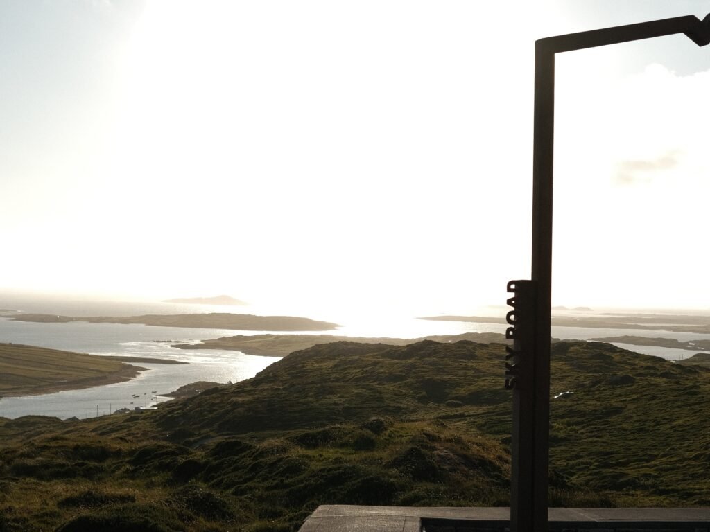 This image highlights a stunning view from the Sky Road in Clifden, part of the Wild Atlantic Way in Connemara, County Galway. The silhouette of a signpost reading "Sky Road" is visible against the backdrop of rolling green hills and the expansive Atlantic Ocean. The distant islands and coastline reflect the evening sunlight, creating a serene and breathtaking scene. This spot perfectly captures the wild, rugged beauty of Ireland's west coast, making it an unforgettable stop for travelers exploring the area by car or foot.