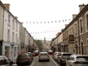 A lively street in Kenmare, County Kerry, Ireland, lined with shops, bistros, and pubs, including The Purple Heather Bistro, with bunting flags hanging above the road.