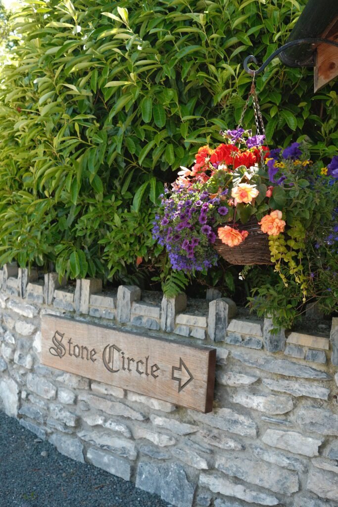 Sign to the Kenmare Stone Circle, Ireland – A rustic wooden sign pointing toward the Kenmare Stone Circle, surrounded by vibrant flowers and greenery. A historic landmark and must-see attraction in Kenmare, County Kerry, Ireland.