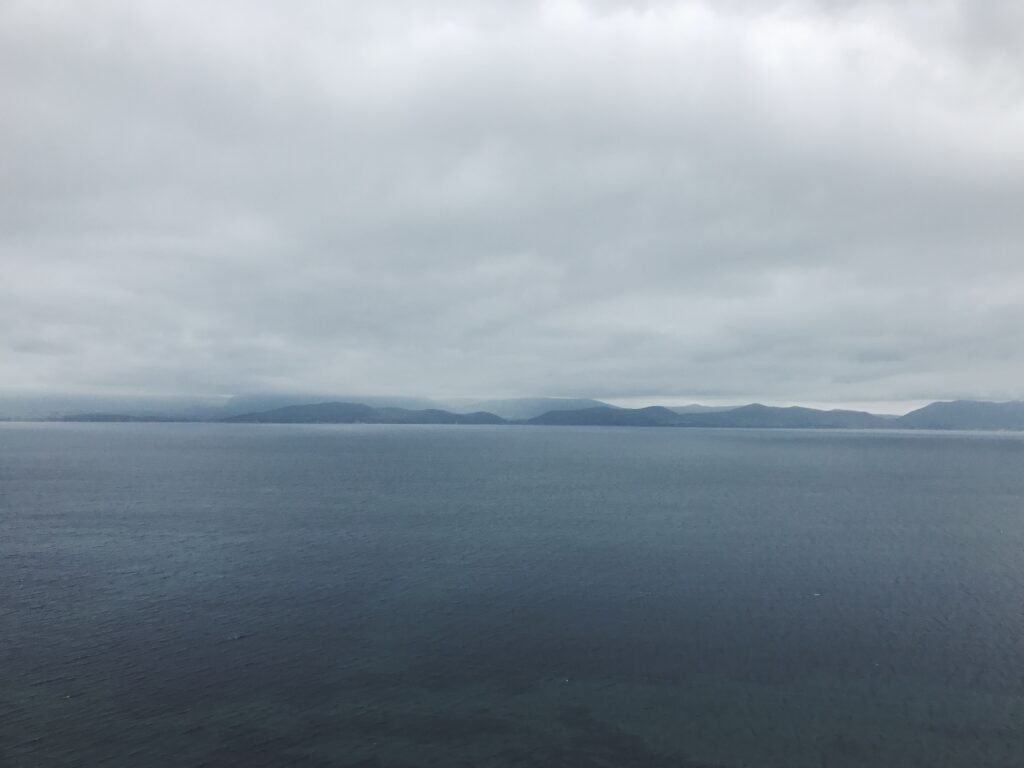 Expansive seascape view along the Ring of Kerry, Ireland, driving route featuring calm blue waters and a distant range of misty mountains under an overcast sky.