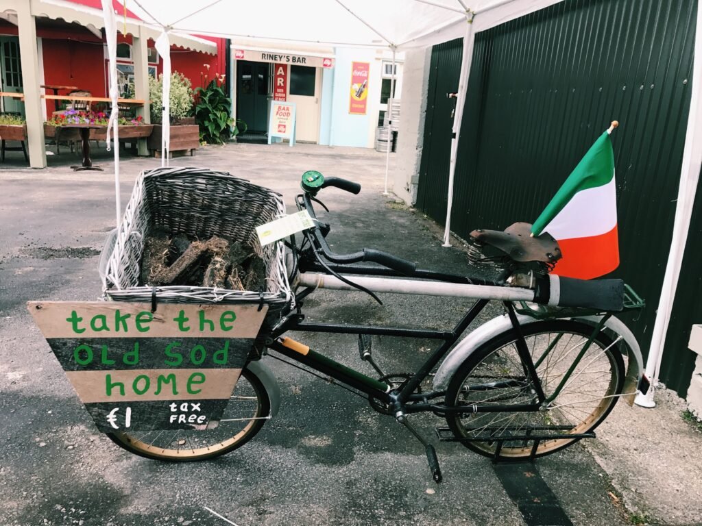 Quirky display in Sneem Village along the Ring of Kerry, Ireland, featuring a vintage bicycle with a basket of turf, an Irish flag, and a humorous sign reading 'Take the Old Sod Home' for €1. A great stop to add to your Ring of Kerry Itinerary.